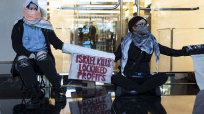 Activists with covered faces and lockboxed arms sit behind a sign reading "ISRAEL KILLS, LOCKHEED PROFITS" during a sit-in outside of the Lockheed Martin headquarters