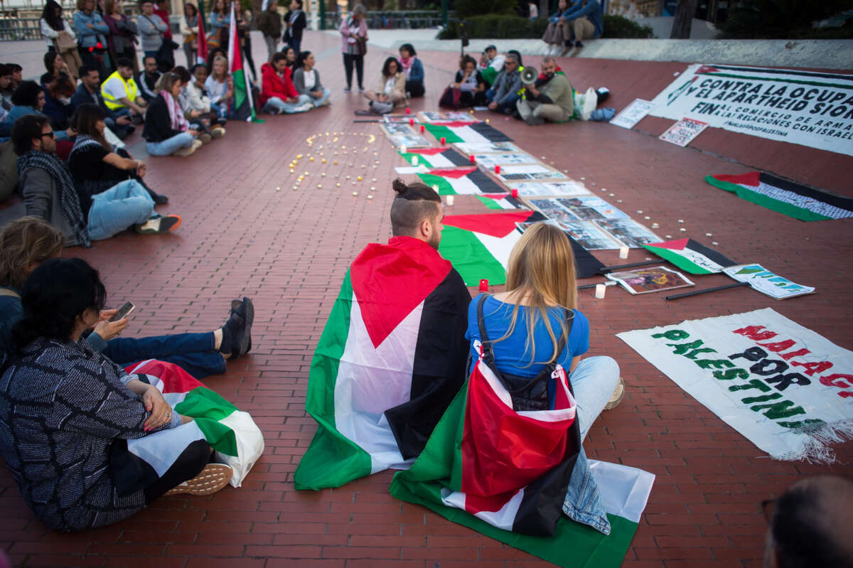 Two people draped in their own Palestinian flags sit with others at an outdoor demonstration