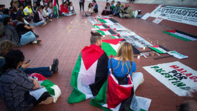 Two people draped in their own Palestinian flags sit with others at an outdoor demonstration