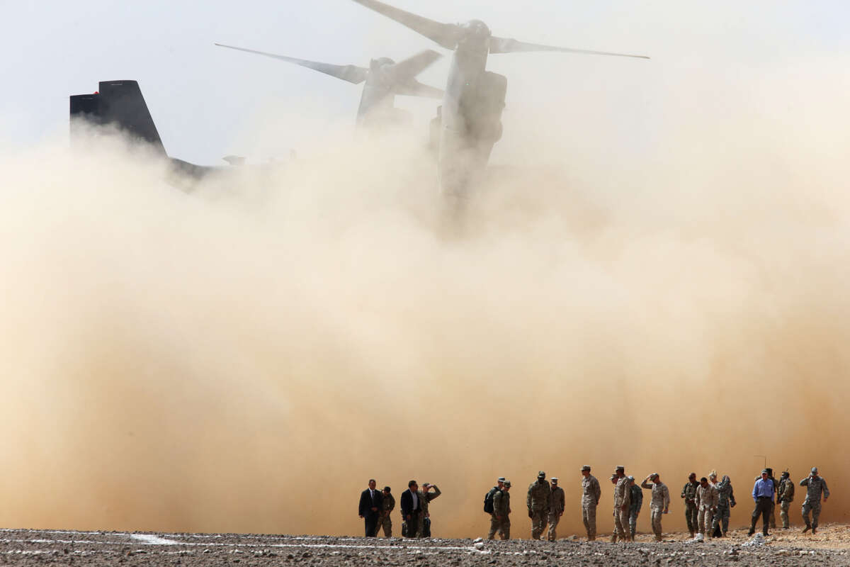 An army aircraft kicks up dirt as troops are seen in the foreground