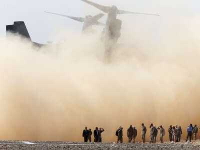 An army aircraft kicks up dirt as troops are seen in the foreground