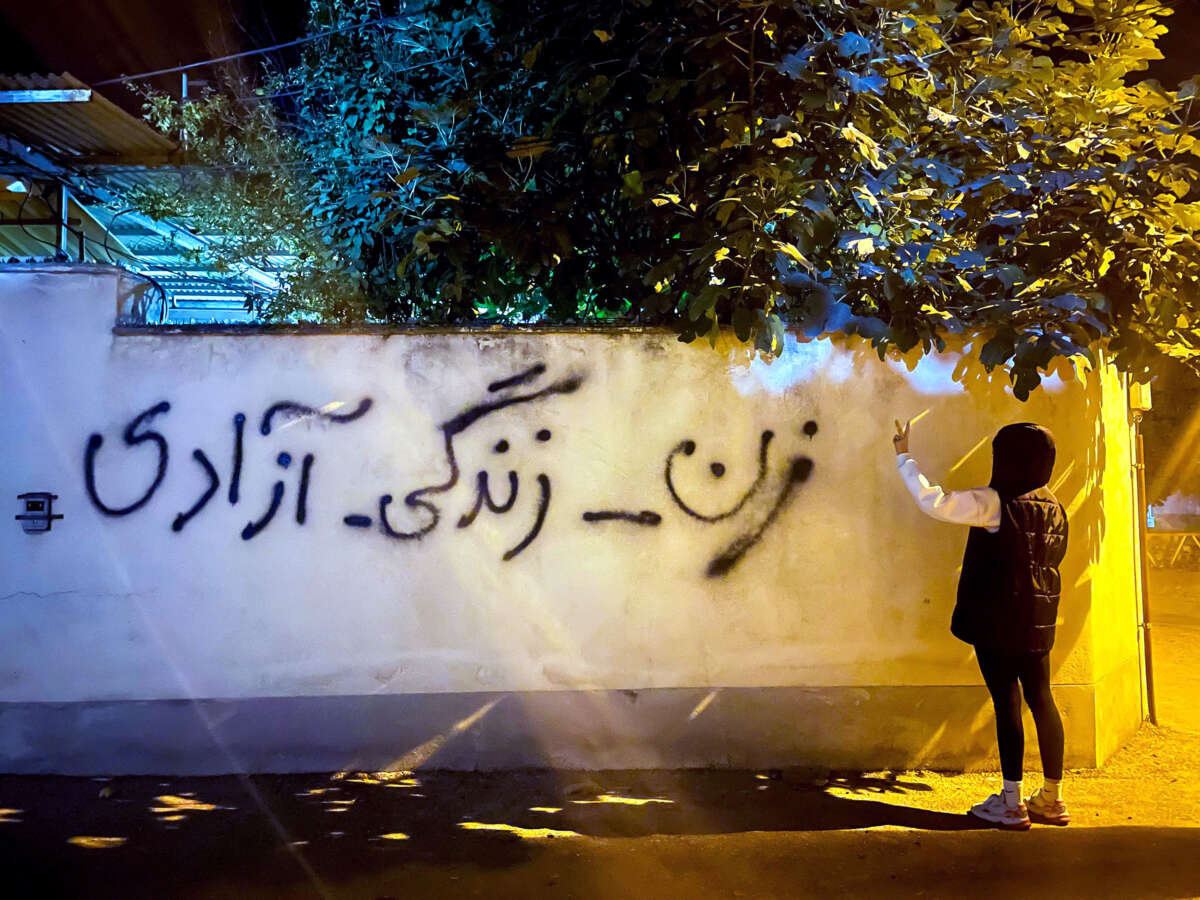 A girl in a black hoodie with her back turned to the camera makes a peace sign while standing beside a painting reading "Women, life, freedom,"