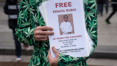 A man in a blazer with cannabis leaf print holds a laminated flyer with a photo of Edwin Rubis, along with text calling for his freedom from his 40-year prison sentence for marijuana