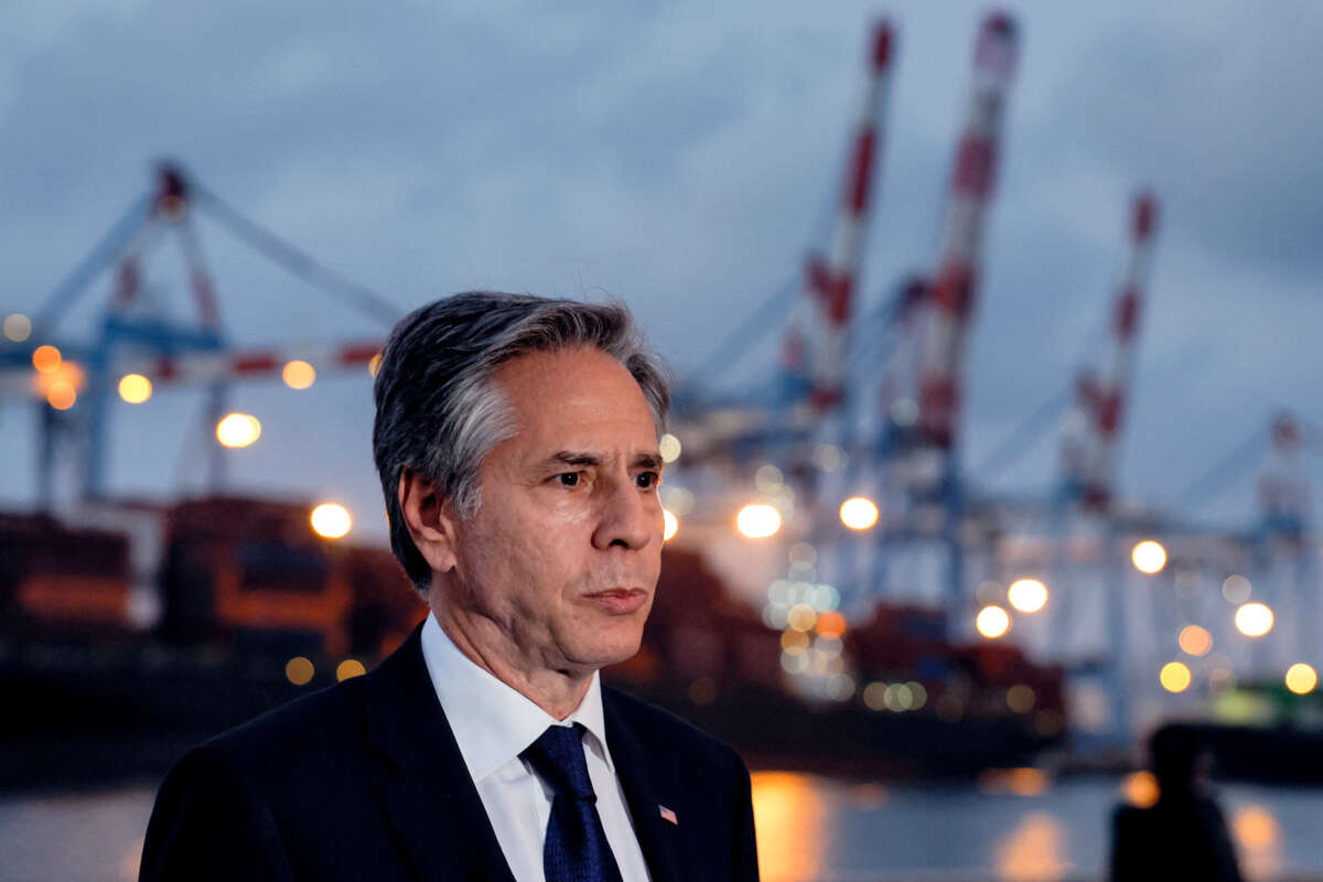 Anthony Blinken stands at a port with cranes and bridges in the background