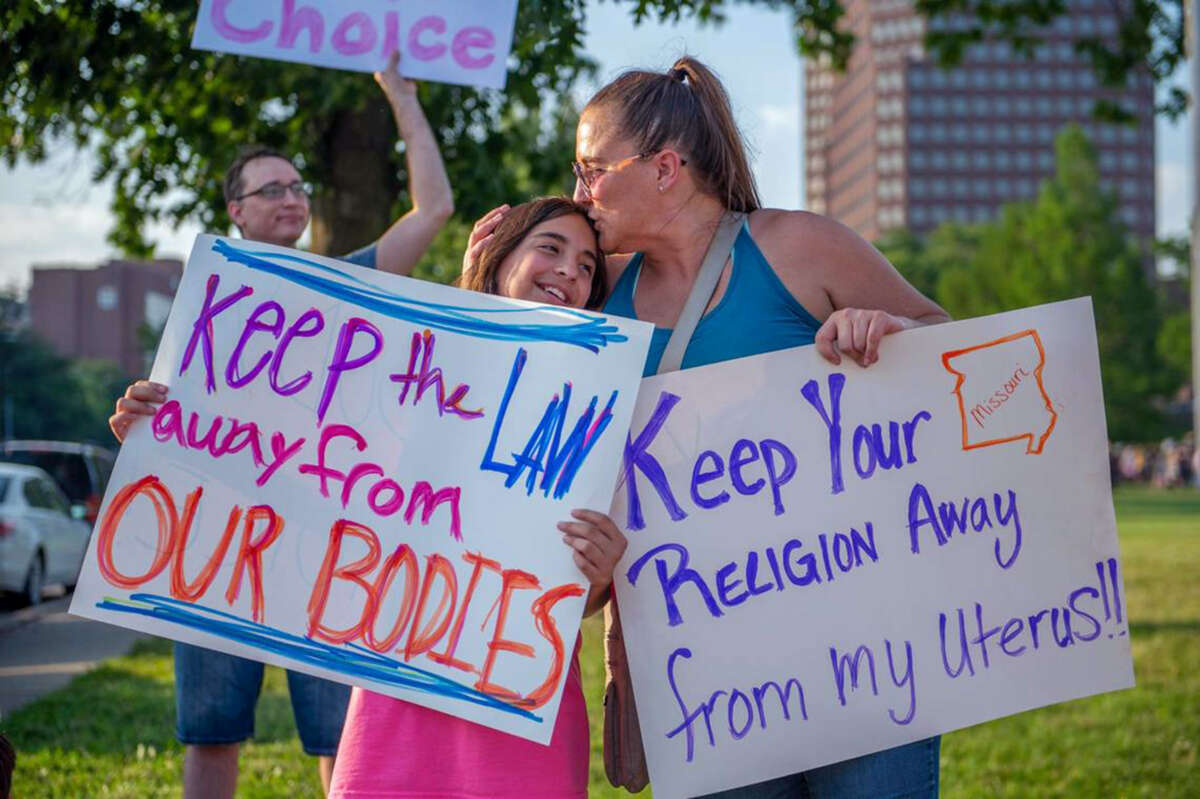 A mother kisses her daughter on the head as they hold signs reading "Keep your religion away from my uterus!" and "Keep the LAW away from OUR BODIES" during an outdoor protest