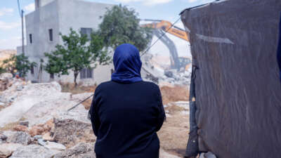 A woman in a blue hijab watches Israeli forces tear down her home