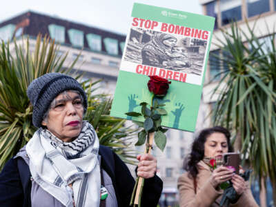 A protester in a kuffiyeh holds a sign reading "STOP BOMBING CHILDREN" during an outdoor protest