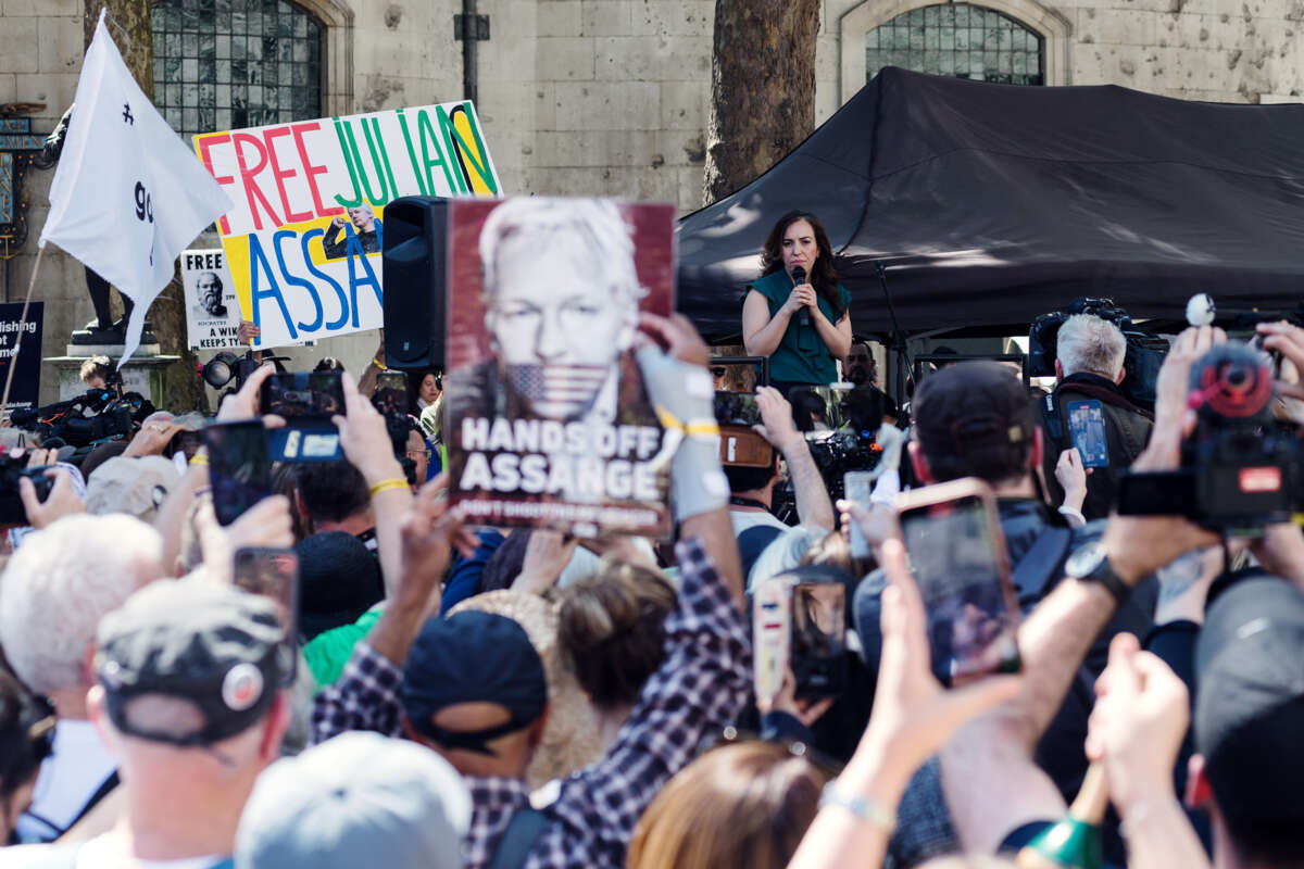 Stella Assange speaks at a podium as people display signs showing her husband, Julian Assange, censored by the united states flag