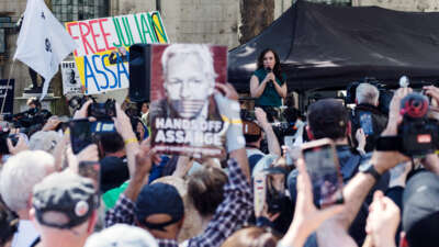 Stella Assange speaks at a podium as people display signs showing her husband, Julian Assange, censored by the united states flag