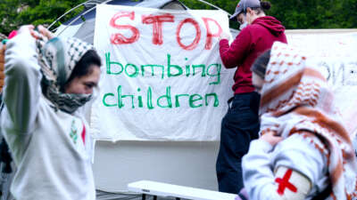 A person affixes a sign reading "STOP BOMBING CHILDREN" to a tent in a campus encampment