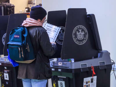 A voter wearing a beanie and a backpack stands at a booth in preparation to vote