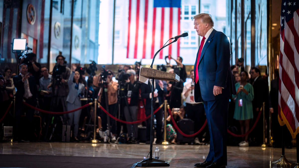 Donald Trump speaks into a microphone in front of a U.S. flag and a throng of reporters