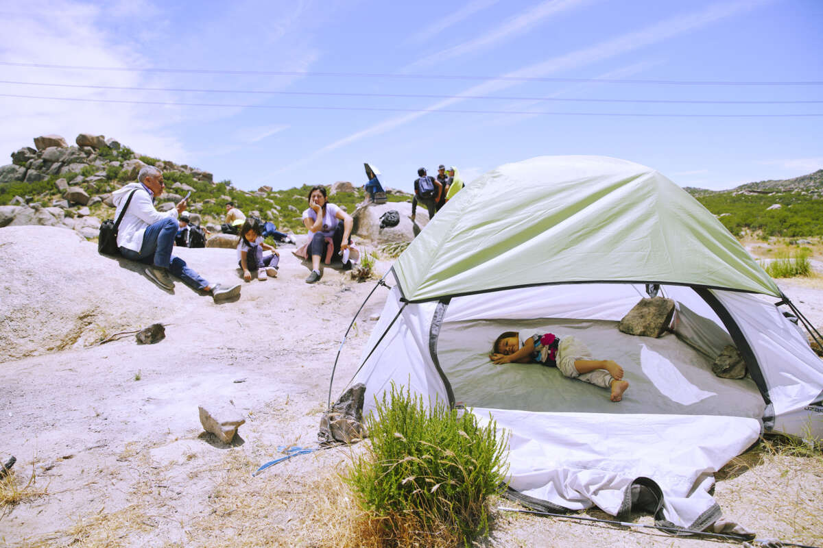 A baby sleeps in a tent in a desert as adults sit nearby