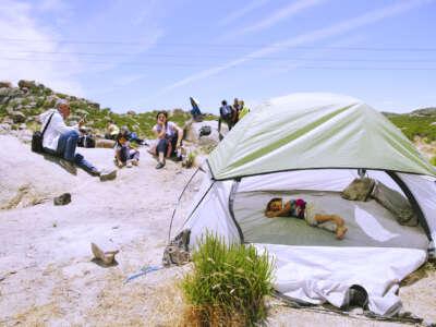 A baby sleeps in a tent in a desert as adults sit nearby