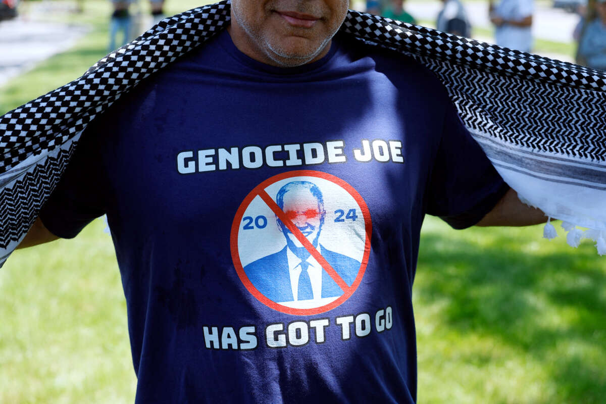 A man moves his keffiyeh aside to show a shirt he's wearing that reads "GENOCIDE JOE HAS GOT TO GO"