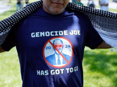 A man moves his keffiyeh aside to show a shirt he's wearing that reads "GENOCIDE JOE HAS GOT TO GO"