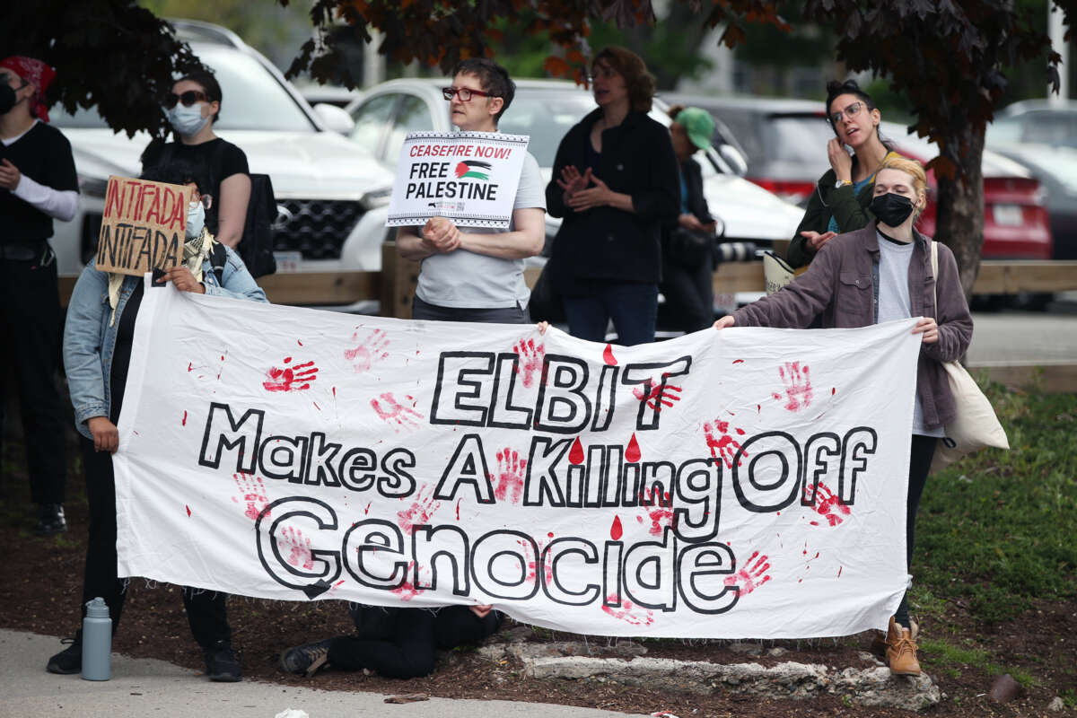 Protesters stand behind a banner reading "ELBIT MAKES A KILLING OFF GENOCIDE" during an outdoor demonstration