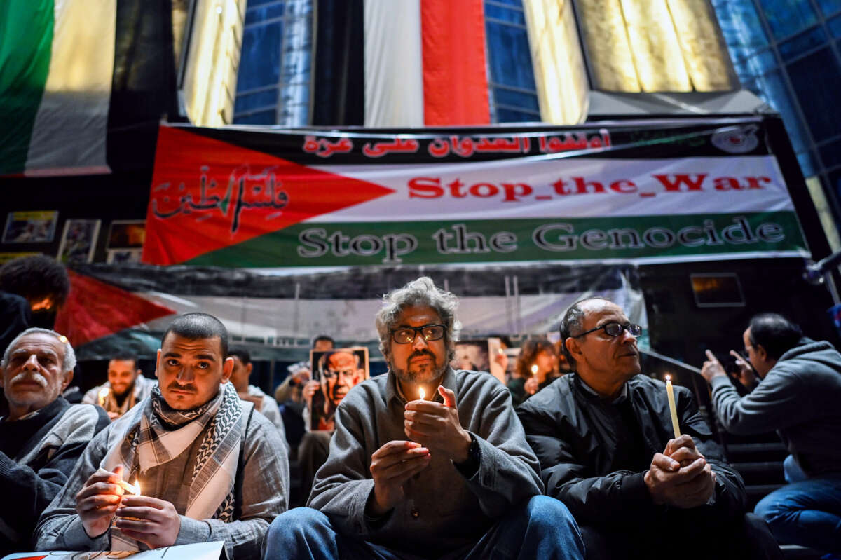 Men holding candles sit in front of a Palestinian flag bearing the words "STOP THE WAR; STOP THE GENOCIDE" during an outdoor vigil
