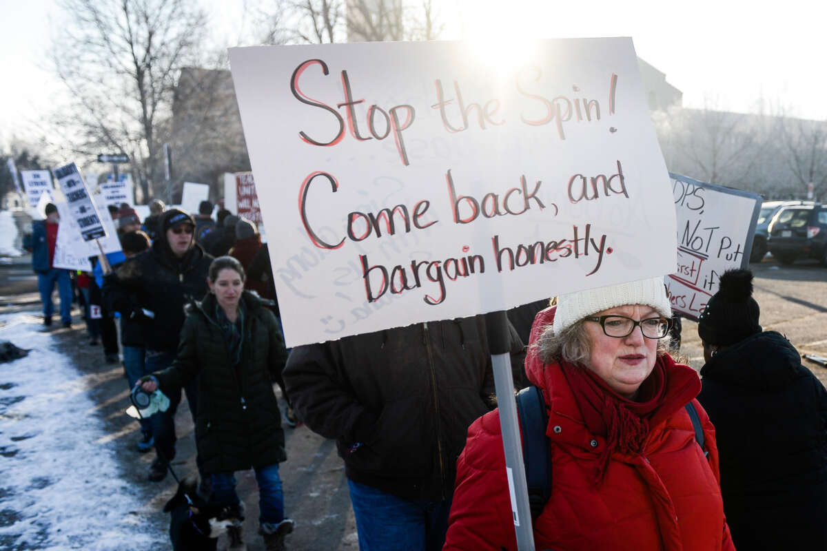 A teacher in a red coat holds a sign reading "STOP THE SPIN; COME BACK, AND BARGAIN HONESTLY" during an outdoor protest