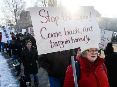 A teacher in a red coat holds a sign reading "STOP THE SPIN; COME BACK, AND BARGAIN HONESTLY" during an outdoor protest