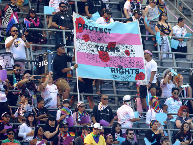 People in bleachers raise a banner reading "PROTECT TRANS RIGHTS" during a sporting event