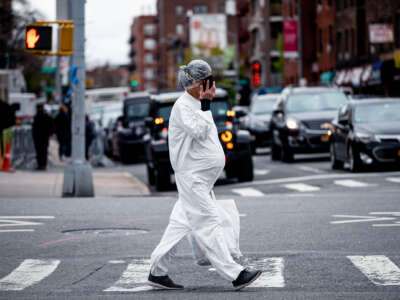A pregnant woman in ppe crosses a busy intersection while talking on her cell phone