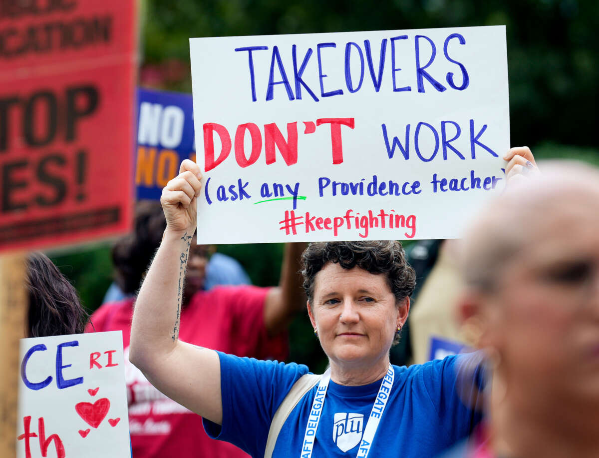 A teacher holds up a sign reading "TAKEOVERS DON'T WORK; (ask any Providence teacher)" during an outdoor protest