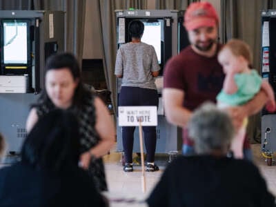 A person votes at a kiosk in the background as others register to vote in the foreground