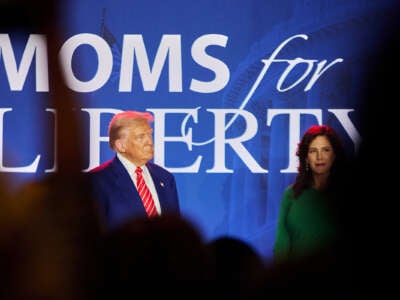 Donald Trump speaks on a stage with the words "MOMS FOR LIBERTY" projected on a screen behind him