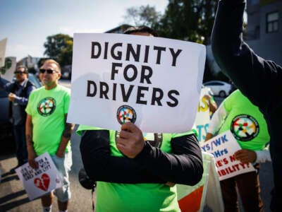 A person in a green shirt holds up a sign reading "DIGNITY FOR DRIVERS" with the Rideshare Drivers United logo beneath the text