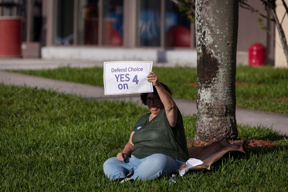 A protester sits on grass while holding a sign reading "DEFEND CHOICE; YES ON 4" during an outdoor protest