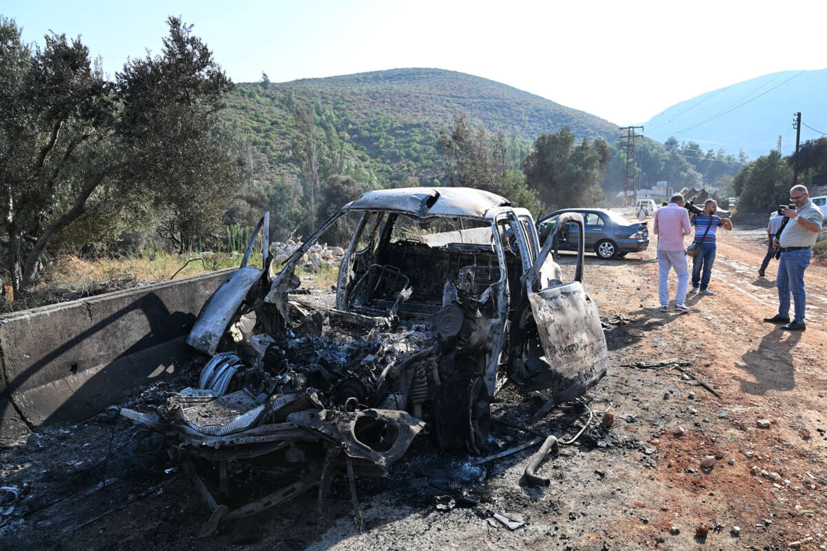 A bombed car sits on the side of the road while people inspect it