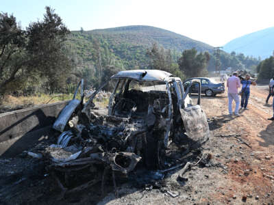 A bombed car sits on the side of the road while people inspect it