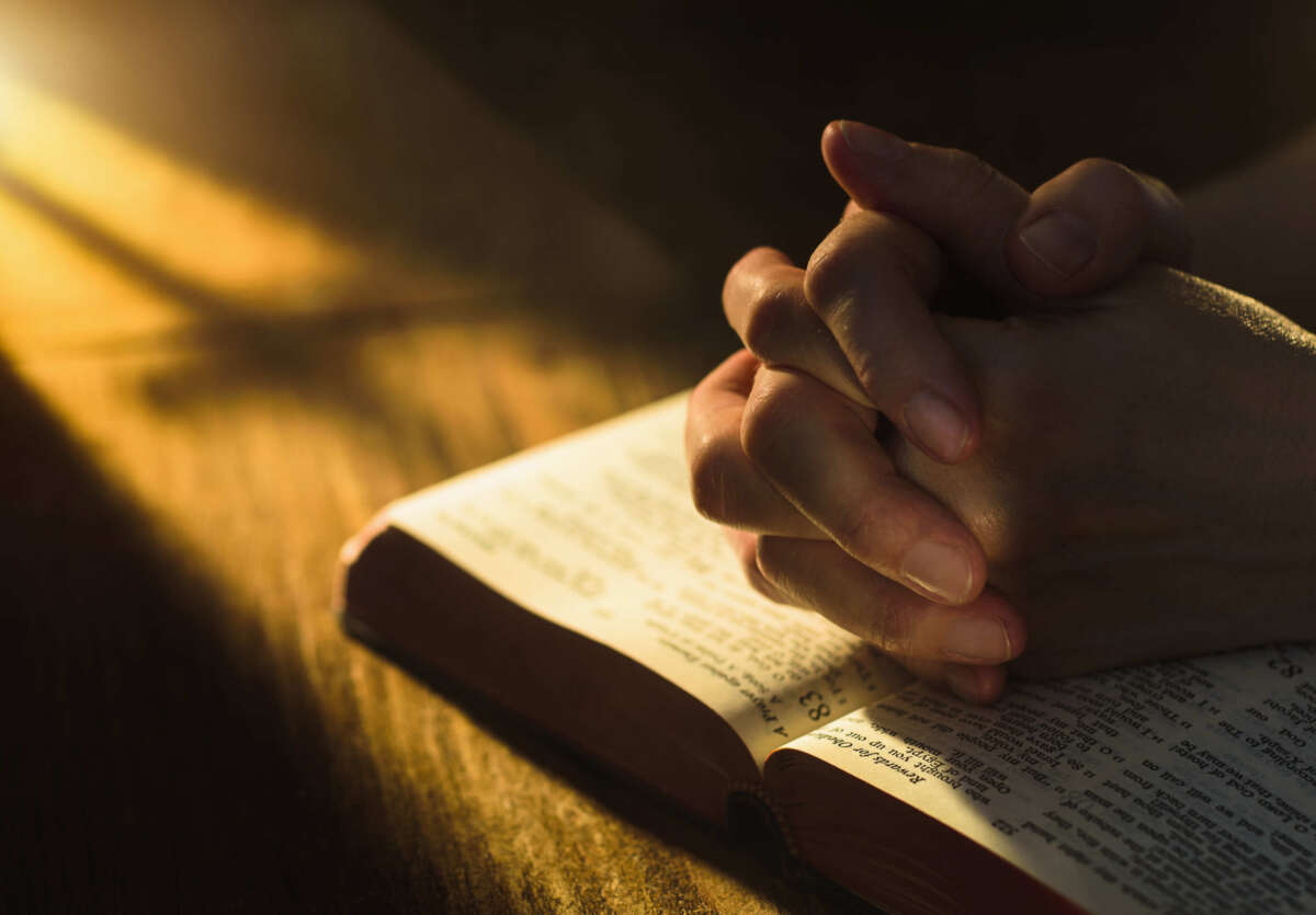 Child's hands clasped in prayer over bible on wooden table with early dawn lighting