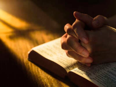 Child's hands clasped in prayer over bible on wooden table with early dawn lighting