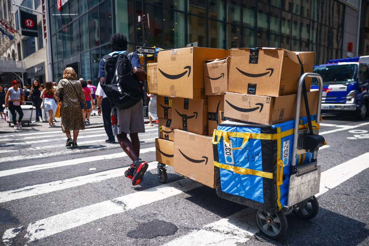 A lone worker drags an overloaded dolly covered in Amazon packages across a busy intersection