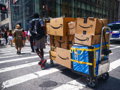 A lone worker drags an overloaded dolly covered in Amazon packages across a busy intersection