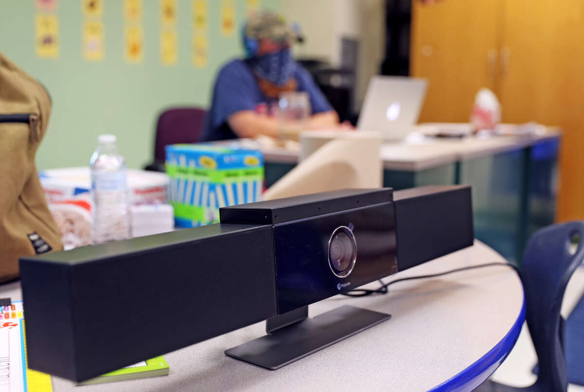 A teacher in ppe is seen out of focus behind a video camera set up on a desk