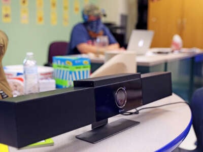 A teacher in ppe is seen out of focus behind a video camera set up on a desk