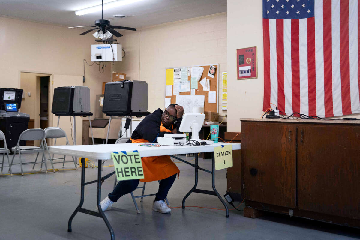 A poll worker, wearied, sits at a table next to an U.S. flag hanging on a nearby wall