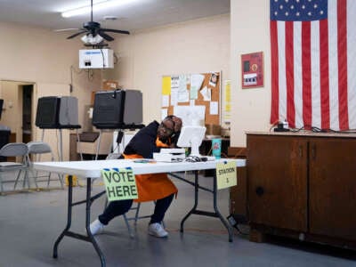 A poll worker, wearied, sits at a table next to an U.S. flag hanging on a nearby wall