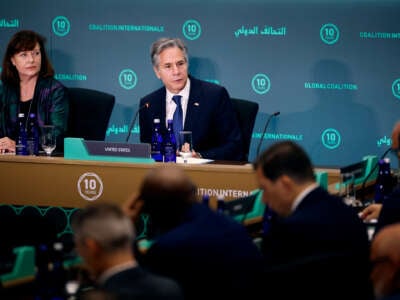 Anthony Blinken sits at a table during a presentation and looks to his left at someone speaking