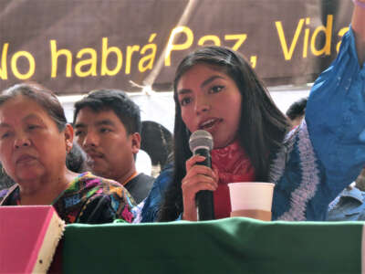 A woman speaks into a microphone while seated beside others at a table