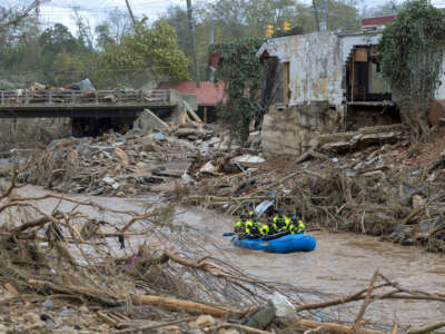 An inflatable raft carrying people floats through floodwaters beside a partially-destroyed building