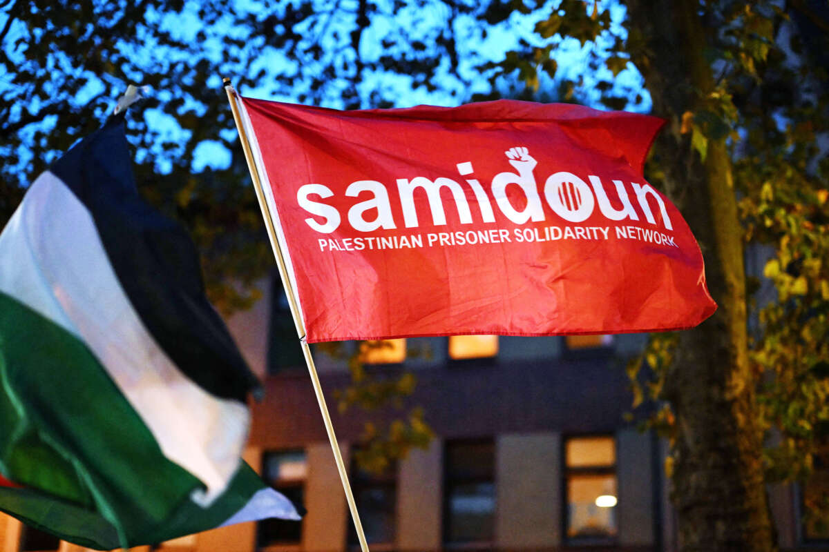 A red flag with the words "Samidoun Palestinian Prisoner Solidarity Network" is raised at an outdoor rally