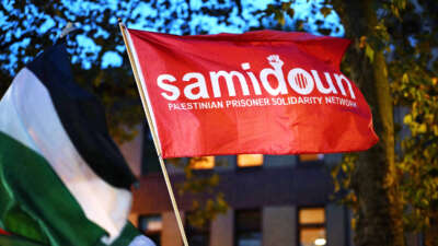 A red flag with the words "Samidoun Palestinian Prisoner Solidarity Network" is raised at an outdoor rally