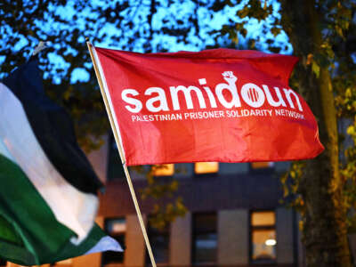 A red flag with the words "Samidoun Palestinian Prisoner Solidarity Network" is raised at an outdoor rally