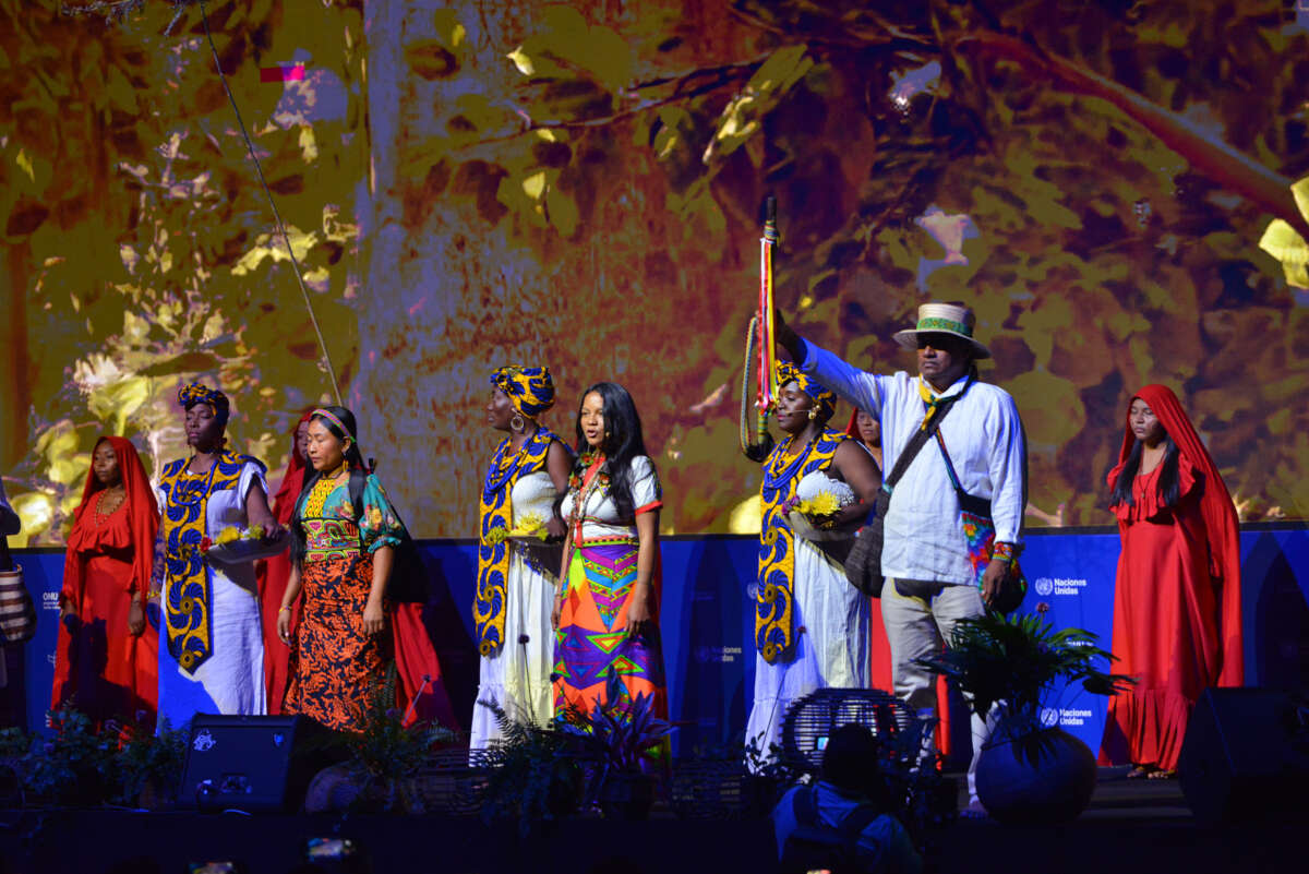 Women dressed robes from various indigenous religious practices stand together on a stage