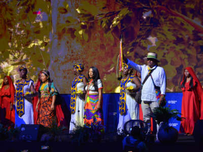 Women dressed robes from various indigenous religious practices stand together on a stage