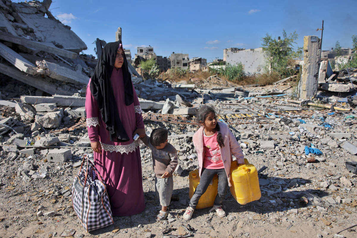 A woman and her two children carry water and a single bag of their belongings amidst destroyed buildings